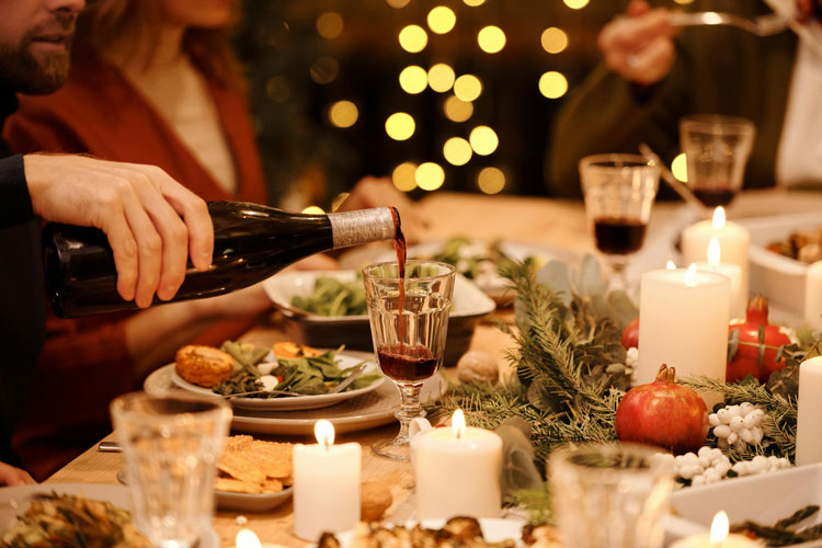 Festlich gedeckter Tisch mit Kerzen, Grün, Granatäpfeln; Person schenkt Rotwein ein, Essen und Gäste sichtbar.