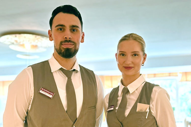 Zwei Personen in Uniform mit weißen Hemden, grauen Westen und Krawatten, Namensschildern und Deckenleuchte im Hintergrund.