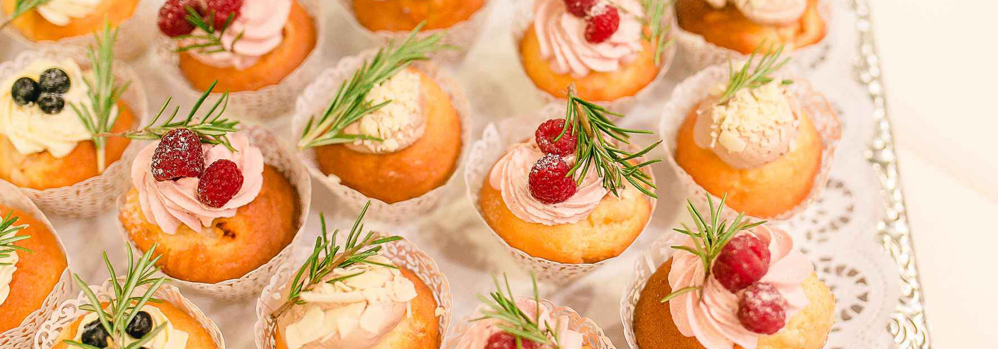 Tablett mit Cupcakes in Papierförmchen, dekoriert mit Beeren, Creme und Rosmarin, auf Spitze angerichtet.